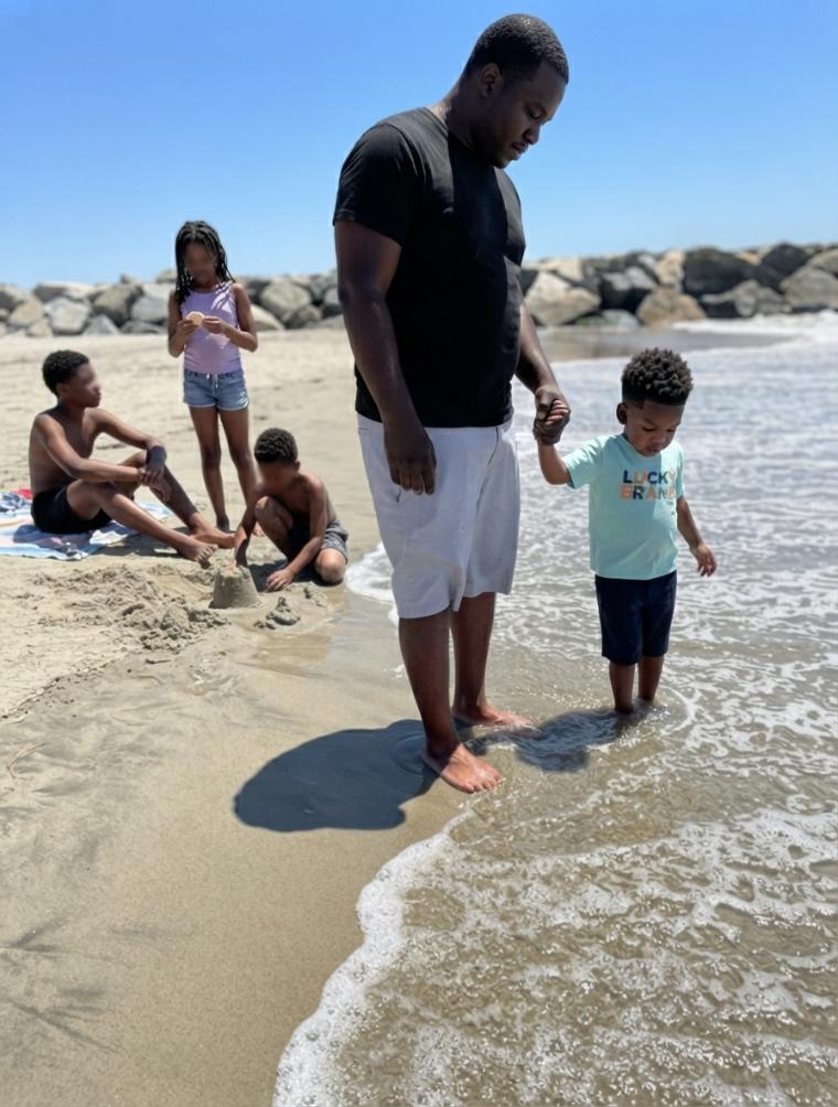 A sandcastle taking shape on the beach while a father steadies his toddler in the shallow water nearby