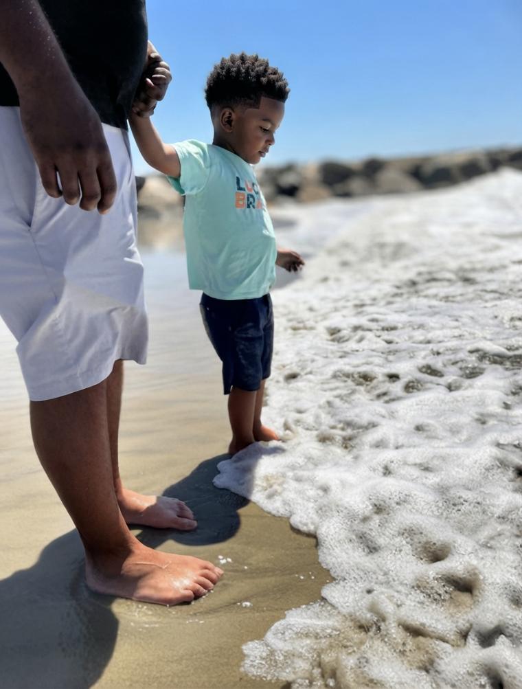 A father and his youngest standing at the edge of the surf, hand in hand, as the foam runs over their feet